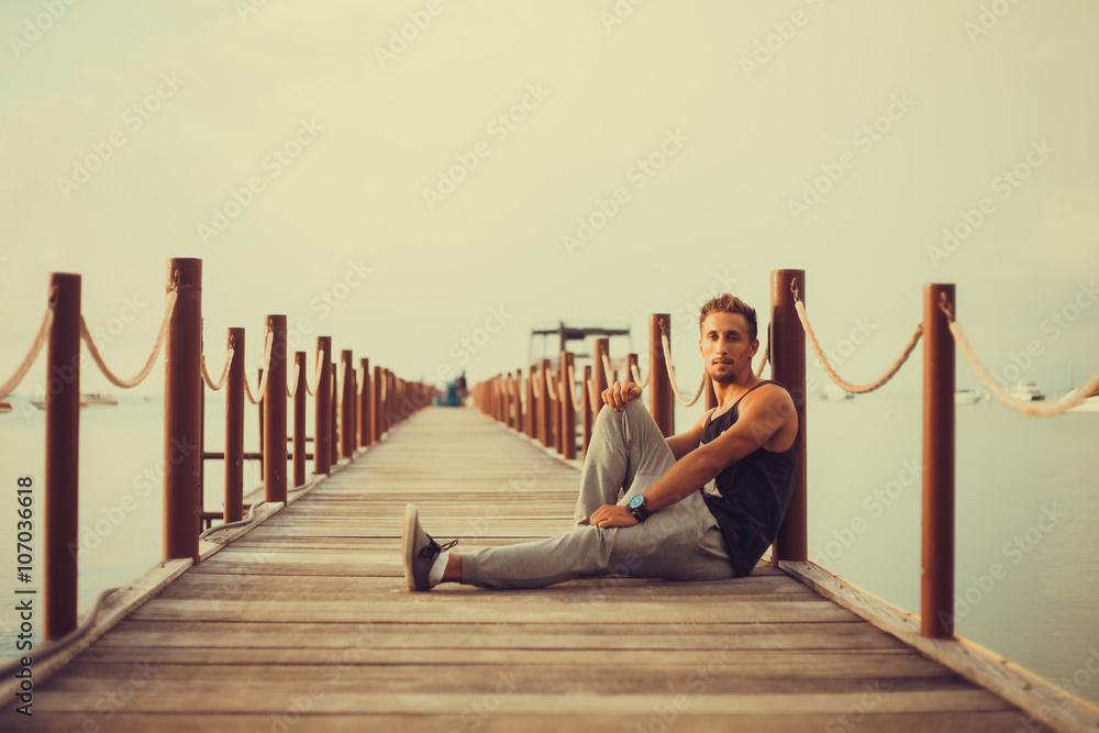 Wall mural young guy with a beard in a t-shirt and sunglasses posing on the pier,Rugged and manly. Confident young bearded man looking at camera and adjusting eyewear while standing outdoors