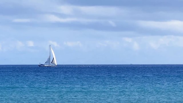 Grey white sailboat crossing the wide blue calm ocean. No waves and a partly cloudy sky.