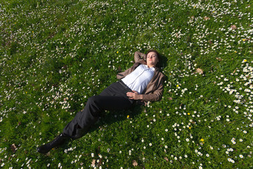 Relaxing young woman in pants and leather jacket lying on grass and spring flowers
