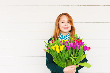 Portrait of redhead girl of 8-9 years old, holding bright bouquet of colorful fresh tulips, standing against white wooden background