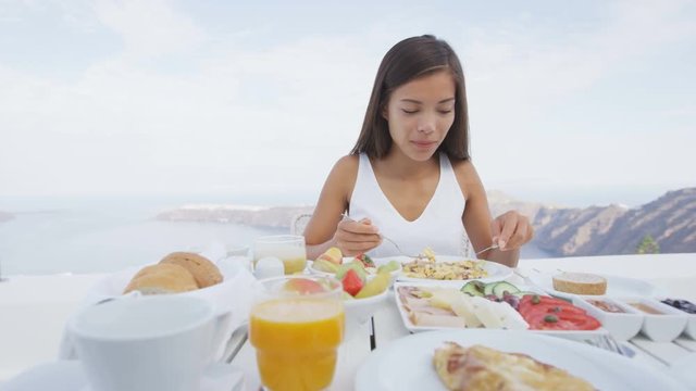 Beautiful tourist eating breakfast on terrace resort. Woman is enjoying tasty healthy food served by sea. She is on her luxury vacation travel holidays on Santorini, Greek Islands, Greece, Europe.