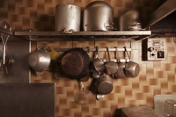 Obraz na płótnie Canvas Kitchen utencils in old vintage professional bakery. Heavy used industrial pans, pots, whisks and more isolated on steel shelf and hangovers on wall with ceramic bricks
