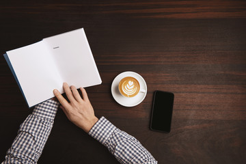 Obraz premium Top view accountant hands in plaid shirt hold opened note book with contents near cup with cappuccino on white ceramic tea plate and phablet phone on red wooden table during coffee break 