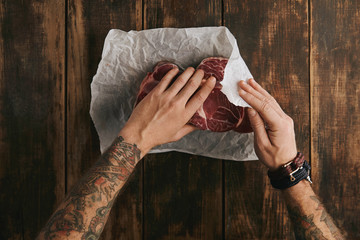 Brutal man with tattooed hands folds three fresh raw meat steaks in white craft paper on vintage...