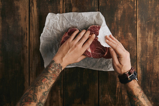 Brutal man with tattooed hands folds three fresh raw meat steaks in white craft paper on vintage aged wooden table, top view, ready to cook
