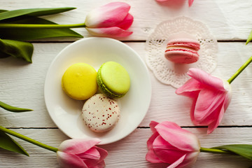 A beautiful flowers pink tulips with colorful macaroons laid on a white platter on white wooden background