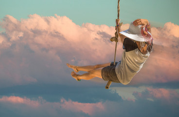 Young woman with shorts, shirt and wide-brimmed hat swinging with sunset sky and cloud background....