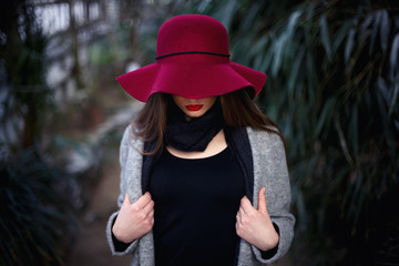 beautiful young brown-haired woman with long hair, beautiful makeup with bright red lips, wearing a gray coat, black pants, a scarf, a hat and sunglasses walking in a greenhouse