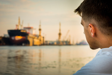 Fototapeta premium Head of the young man sitting on the bank of the river with a blurred ship in the background