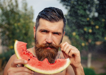 bearded man without clothes with a big juicy ripe watermelon in hands on a background of flowering garden illuminated by bright sunshine