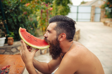 bearded man without clothes with a big juicy ripe watermelon in hands on a background of flowering...