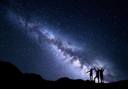 Landscape With Blue Milky Way. Night Starry Sky With Silhouette Of A Happy Family With Raised-up Arms On The Mountain. Beautiful Universe. Space Background