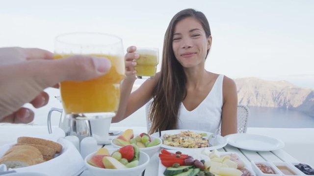 Couple eating breakfast. Happy tourist woman with man toasting juice glasses on terrace resort. Healthy and delicious food served for breakfast. Lady enjoying healthy drink in Santorini, Greece.