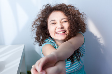 Fototapeta premium Portrait of a beautiful young girl with brown curly hair. Fun and joy. Happy girl smiling and squinting in the morning sun. Delicate blue dress.