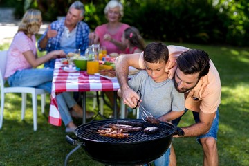 Father teaching son cooking on barbecue with family in background