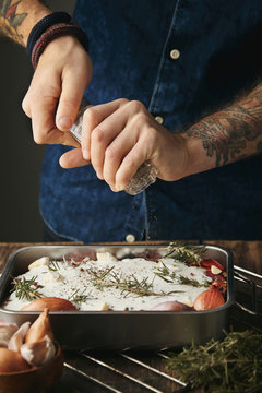 Man with tattooed hands pour pepper  to metallic stainer with three raw meat steaks in greek tzatziki sause. Spices and herbs near: garlic, rosemary, onions. Everything ready to cook 