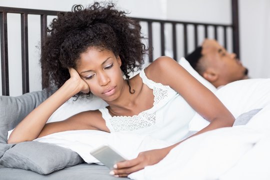 Young Woman Using Mobile Phone While Man Sleeping On Bed