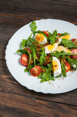 Salad with arugula, cherry tomatoes, eegs and cheese on white ceramic plate over rustic wood background, top view