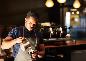 Modern coffee shop with African barista pouring milk into cappuc - obrazy, fototapety, plakaty