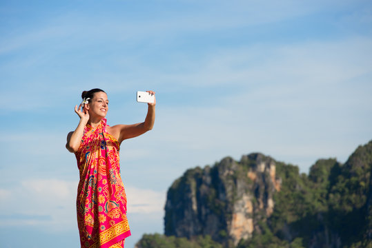 Woman on travel vacation to Krabi, Thailand, taking selfie photo with smartphone camera at the beach. Beautiful brunette model wearing thai summer dress.
