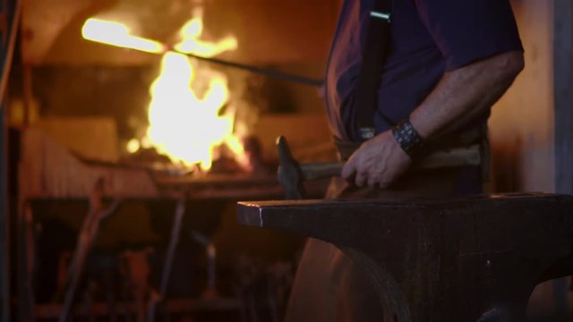 Blacksmith brings dripping hot metal from furnace and creates a shower of sparks as he beats a piece of white hot metal with a hammer on an anvil.  Close up recorded at 180fps.