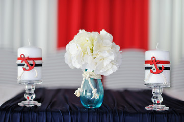На столе стоят две белых свечи с красным декором и ваза с белыми цветами