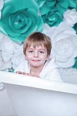 little cute 4-years boy with funny face in bathrobe sit in the bath and smile with white and turquoise flowers on the background