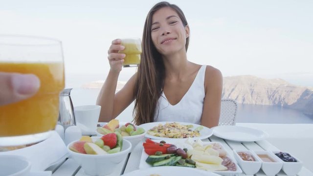 Couple eating breakfast. Smiling tourist woman with man toasting juice glasses on terrace resort. Healthy and delicious food served for breakfast. She is enjoying healthy drink in Santorini.