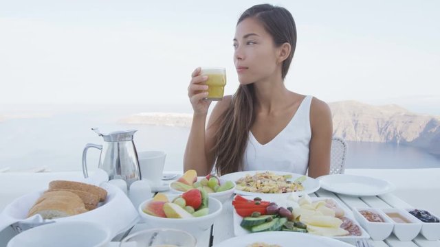 Asian woman eating breakfast. Female tourist having glass of juice on terrace resort. Healthy breakfast served by sea. Elegant Lady on summer vacation in Santorini, Greece, Europe.