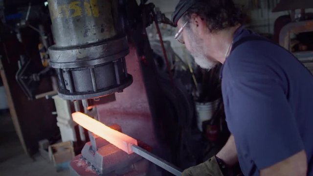 Hot rod of metal being worked by a blacksmith with a large industrial hammer.  Hand held, over the shoulder view, originally recorded in 4K at 60fps.