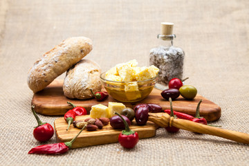 Composition with olive wood, cheese pieces in olive oil, bread and hot peppers with burlap texture in the background