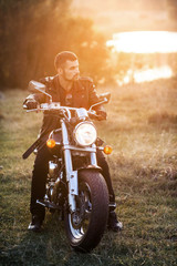 Obraz premium young brutal man in a black jacket and glasses sits near a motorcycle.