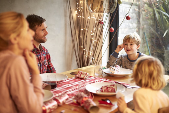 A Family Of Four People, Two Adults And Two Children Seated Around  A Table At Christmas Time, Pulling Crackers. 