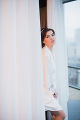 Portrait of a beautiful young girl in a white robe in the bedroom near the window, morning, boudoir, relaxation, evening