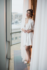 Fototapeta na wymiar Portrait of a beautiful young girl in a white robe in the bedroom near the window, morning, boudoir, relaxation, evening
