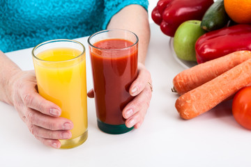 Hands of senior woman putting two glasses with orange and tomato juice on the table near vegetables and fruits
