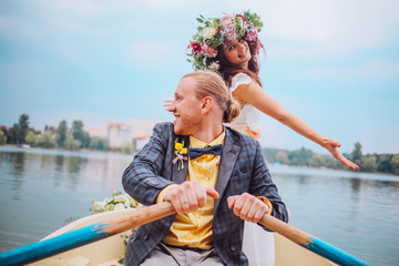 Fototapeta na wymiar bride and groom sitting in an orange row boat floating out of the tree covering and in to the lake. Bride with wreath of flowers