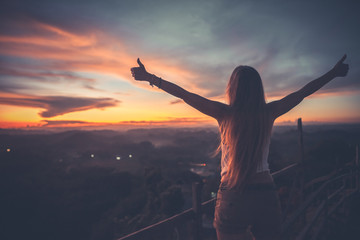 Silhouette of the woman spreading arms with her thumbs up, standing high on the viewpoint with...