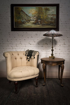 Vintage still life of retro beige armchair, table lamp on antique table and hanged framed painting on dark brown wooden floor and white bricks wall in studio