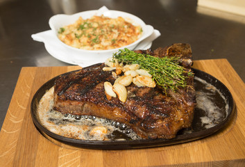 A grilledT Bone steak, roasted garlic and rosemary on a metal plate, served with Macaroni and cheese.