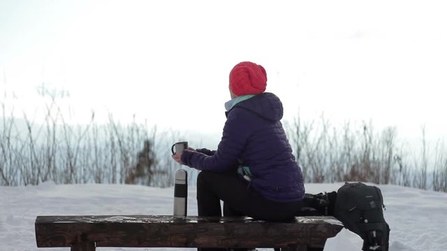 Young woman sitting on a bench and drinking tea from a thermos cup, rear view