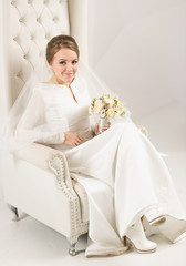 Fototapeta na wymiar Elegant bride in long dress posing on white leather armchair