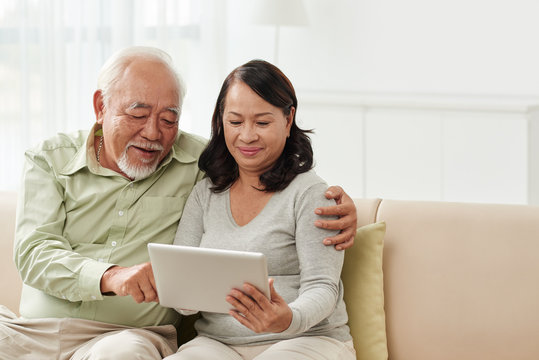 Senior Vietnamese couple with digital tablet sitting on sofa at home