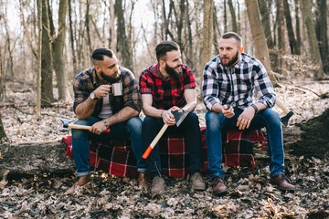 Three bearded men smoke, drink coffee and relax after work in the forest