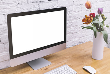 Side view of a computer monitor with a blank screen on a white background of a brick wall and a keyboard on a wooden table with a white vase and fresh flowers, blank for your text or content or design