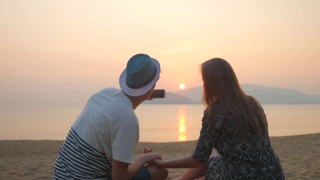 Couple sitting on beach taking photo with smart phone at sunrise. Romantic couple taking pictures using smartphone in summer. Woman and man on holidays travel vacation