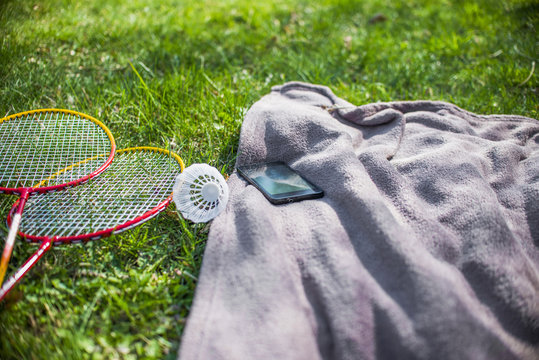 Shuttlecock And Badminton Racket, Mobilephone And Blue Picnic Blanket On The Grass Field