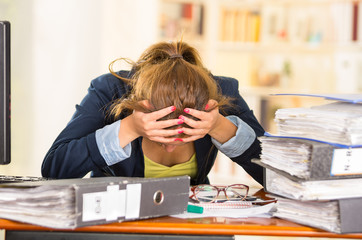 Business woman sitting by desk, paper files spread out, elbows on table and head bent over as...