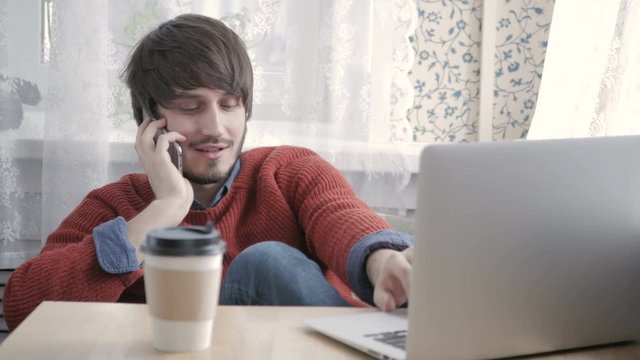 Happy Young Man Freelancer Working with Modern Laptop and Phone in Cafe taking Cup of Coffee