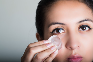 Gorgeous brunette model rubbing ice cube against cheek underneath eye, grey background